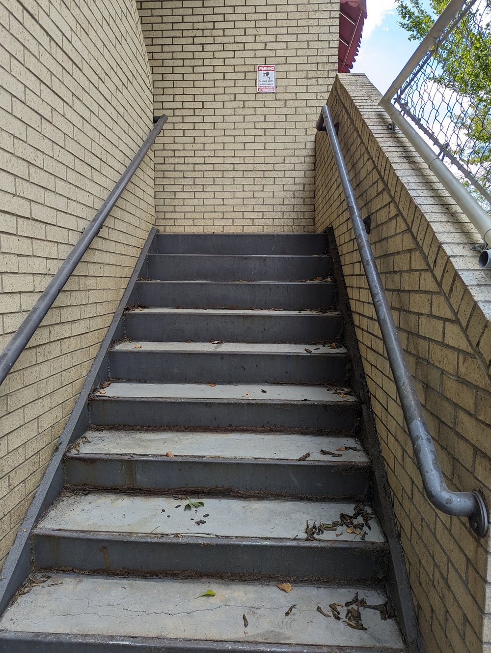 Outdoor metal stairs with black steps rising between brick walls.